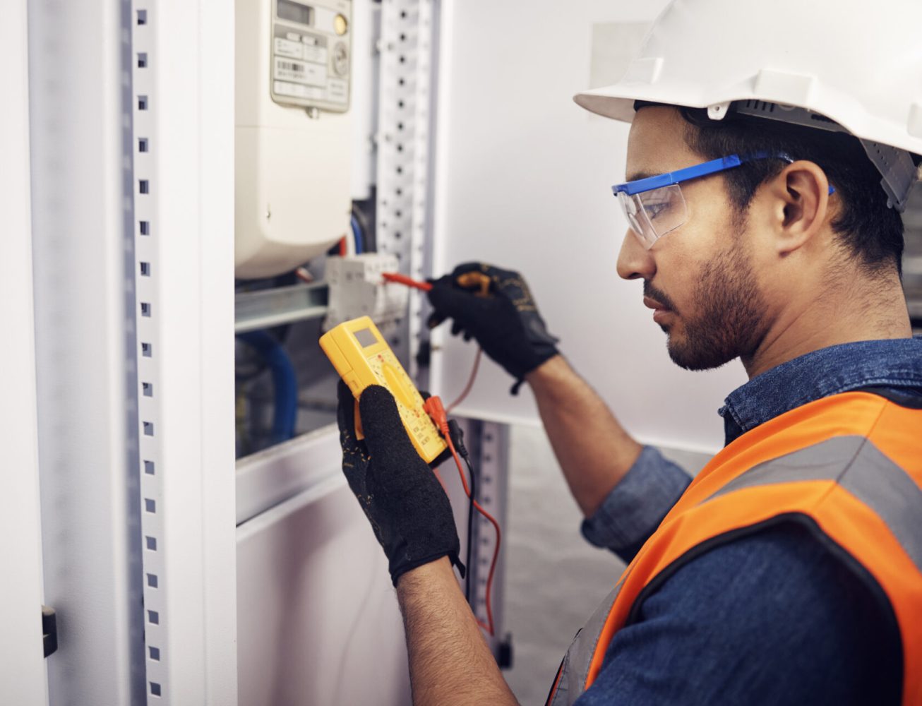 A technician wearing an orange high vis using a multimeter to test a fuse box
